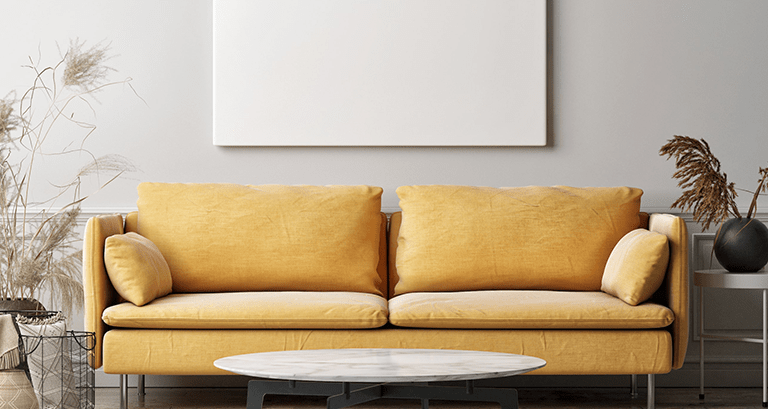 A mustard yellow sofa with two cushions sits against a light gray wall, creating a calming space ideal for therapy or addressing anxiety. Above it hangs a blank white canvas. A round white coffee table and dried plants in vases are in the foreground.