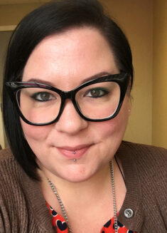 Woman with short dark hair, black cat-eye glasses, and a double lip piercing smiles at the camera. She is wearing a brown cardigan and a red top with a heart pattern, in an indoor setting with tan walls—an inviting presence that reflects her passion for psychotherapy and supporting others through challenges like depression.