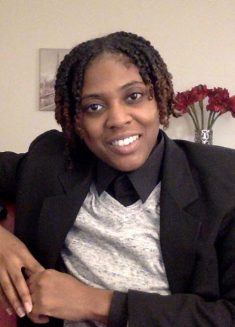 Person with short, curly hair wearing a black blazer, gray sweater, and black shirt smiles while sitting indoors. Red flowers in a vase and wall art are visible in the background, creating a warm atmosphere that suggests a welcoming space for therapy or psychotherapy.