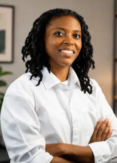 A person with medium-length curly hair, wearing a white collared shirt, smiles confidently with arms crossed in a modern, softly lit office setting—perfectly embodying the professional warmth and approachability of Shamari Saleem, LPC, MS.