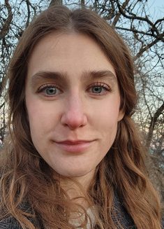 A woman with wavy brown hair stands outside in front of leafless trees, looking into the camera with a slight smile. She wears a dark jacket, and the sky behind her is pale, possibly at sunset. Her calm expression suggests resilience in the face of anxiety or depression.
