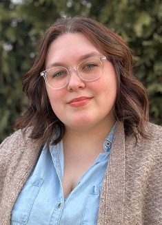 A woman with wavy brown hair and glasses smiles gently at the camera. She is wearing a light blue shirt and a beige cardigan. Green trees are blurred in the background, creating a calm atmosphere that evokes themes of psychotherapy and support for those experiencing depression or anxiety.
