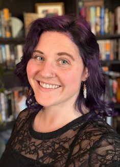 Woman with wavy purple hair and a black lace top smiles in front of a bookshelf filled with books. She has a nose ring and dangling earrings, radiating warmth and openness—qualities that can foster trust in psychotherapy for those experiencing depression or anxiety.