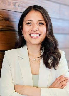 Woman with long dark hair wearing a light-colored blazer and necklace, smiling with arms crossed, standing in front of a wooden wall—embodying confidence and warmth often found in psychotherapy for anxiety or depression.