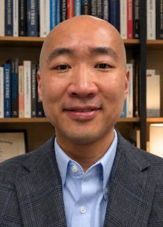A person in a checked blazer and blue shirt smiles in front of a bookshelf filled with books, plants, and a framed certificate, creating an inviting atmosphere often associated with therapy or psychotherapy sessions.