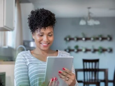 A woman with curly hair and a striped shirt smiles while using a tablet in a modern kitchen with shelves of small plants in the background, perhaps browsing resources about therapy or psychotherapy.