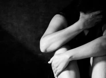 A person sits with knees drawn up, covering part of their face with one hand—a pose that conveys depression or anxiety. The black-and-white image features their body partially illuminated, while the background remains in shadow.