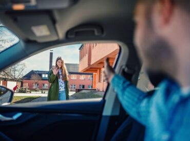 A person inside a car waves to a smiling woman standing outside near a building. She is waving back, and the scene appears friendly and welcoming—conveying a sense of connection often fostered in therapy. The photo is taken from inside the car.