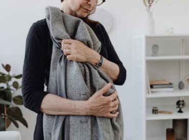 An older woman wearing glasses gently hugs a gray coat, standing in a bright room with shelves and plants in the background, perhaps finding comfort during therapy or moments of anxiety.
