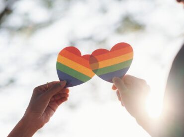 Two hands each hold a paper heart decorated with rainbow stripes, symbolizing LGBTQ+ pride and the importance of therapy in supporting those facing depression or anxiety, against a bright, sunlit background.