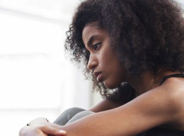 A young woman with curly hair sits indoors, looking pensive and thoughtful, her head resting on her knees—a quiet moment that reflects her experience with anxiety and the journey toward healing through therapy.