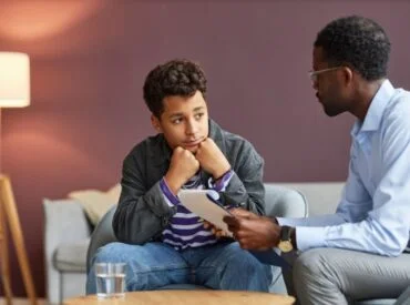 A teenage boy sits with his chin on his hands, looking thoughtful, while an adult man holding a clipboard talks to him in a cozy room during a therapy session for depression and anxiety.