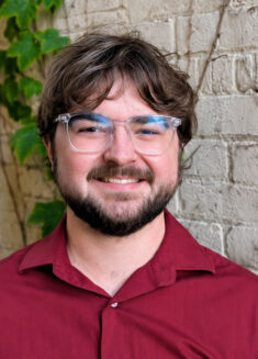 Nick Schiller, a person with brown hair, a beard, and glasses, smiles while standing in front of a white brick wall with green leaves growing on it. He is wearing a maroon shirt.